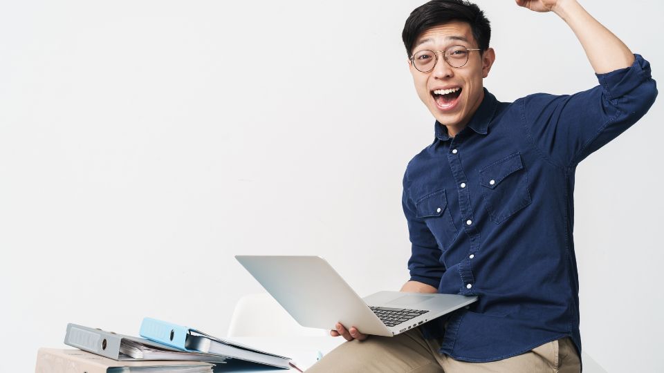 Egy fiatal férfi örömmel ül egy széken, laptopot használva, boldogan integet.