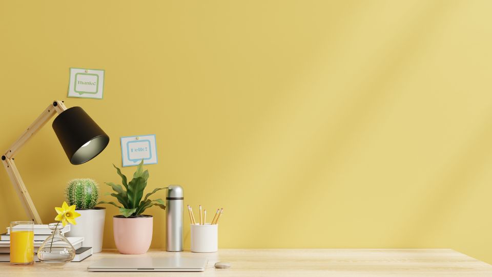 Egy világos sárga fal előtt elhelyezett íróasztal, rajta növények, lámpa és egyéb irodai kellékek.