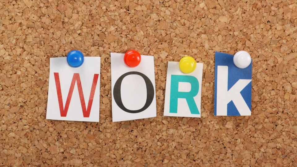 A falitáblán színes papírokon a 'WORK' felirat látható, különböző színű gombostűkkel rögzítve.