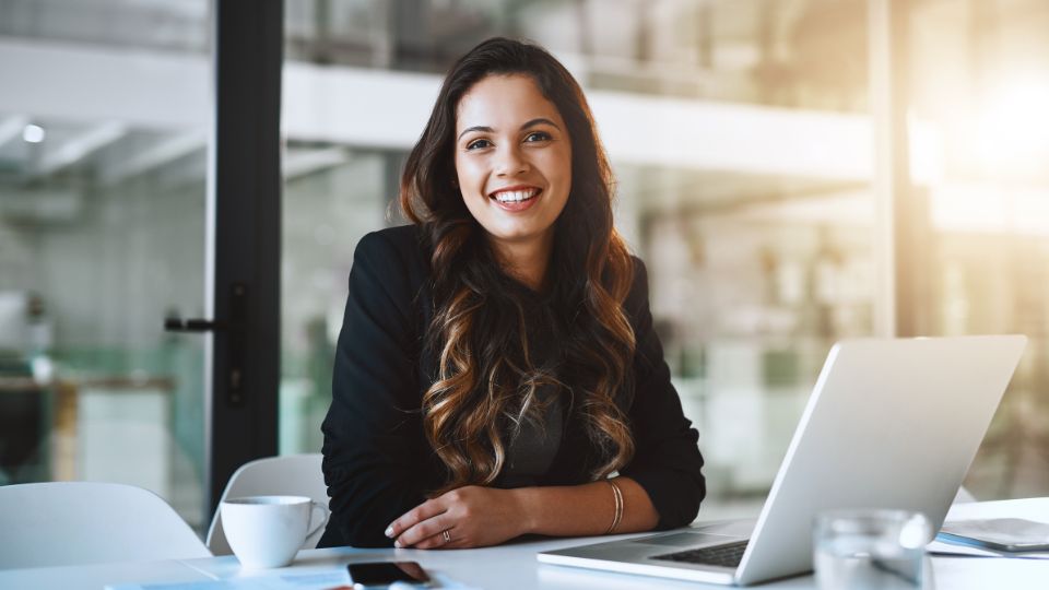 Egy fiatal nő mosolyogva ül az asztalnál, laptop és kávé mellett.