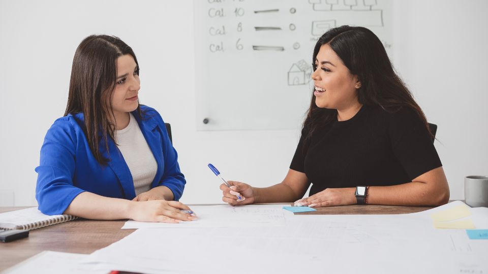 Két nő beszélget egy asztalnál, jegyzetek és tervek között, barátságos légkörben.
