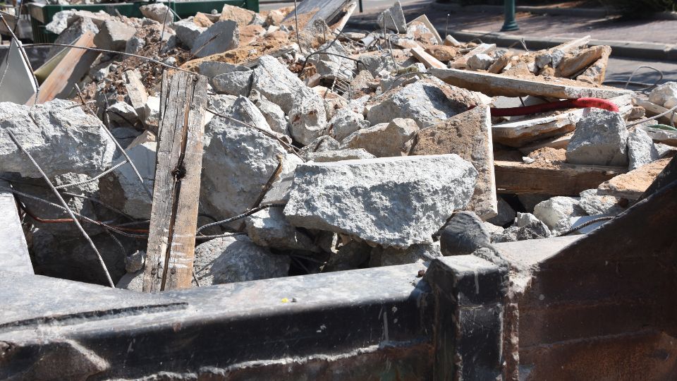 Egy konténer tele építési törmelékkel, kövek és fadarabok láthatók benne.
