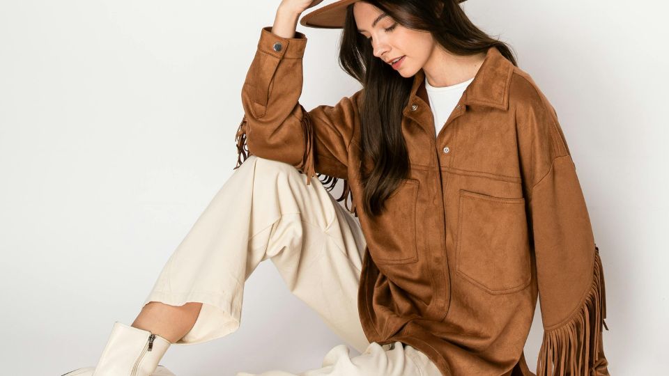 Egy fiatal nő barna francia kabátban és kalapban ül, fehér nadrágban.