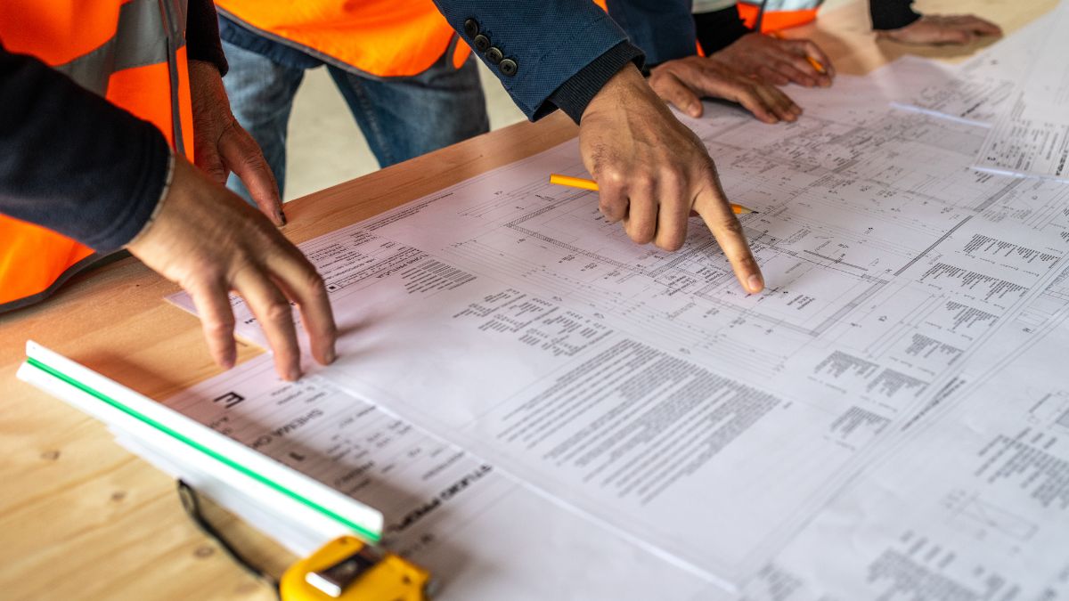Két ember építési terveket néz, egy asztalon elterítve, figyelmesen elemzik a részleteket.